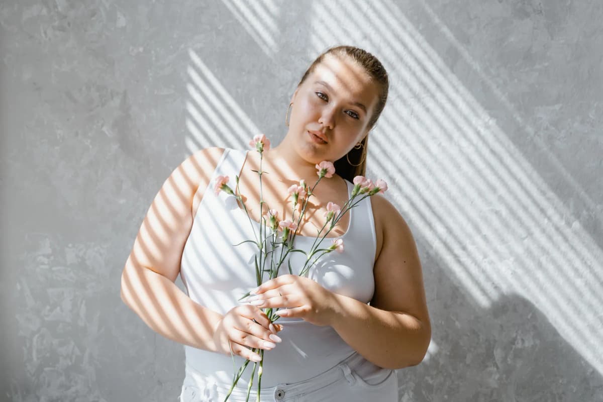 Plus-size woman in tank top holding flowers with sunlight filtering through blinds.